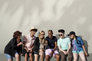 A diverse group of six young people sits together and looks at one of their smartphones, as if watching a video. They are all laughing and happy.